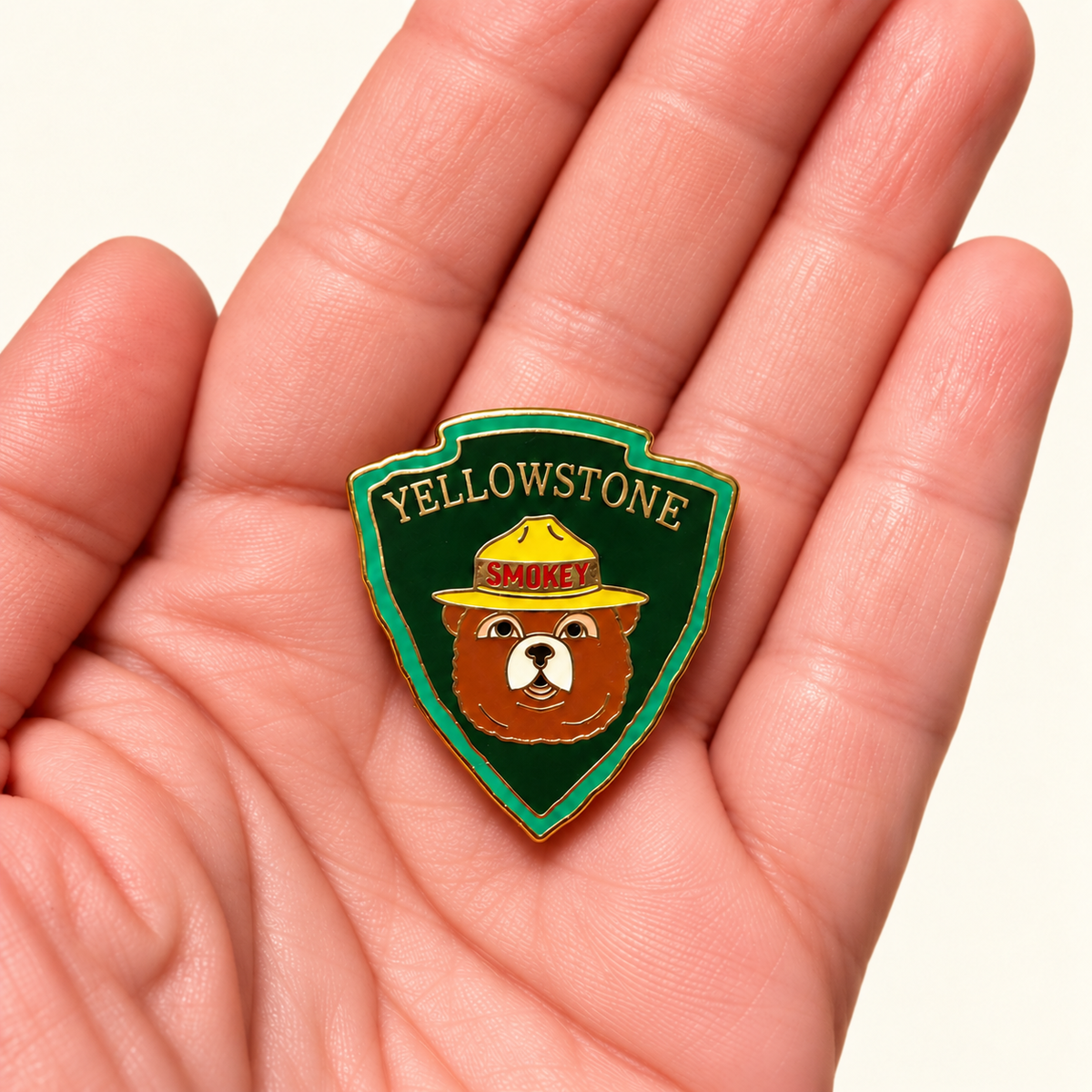 A small enamel pin shaped like a bear badge with the words 'YELLOWSTONE' and 'SPROKER' is held in the center of an open hand. The pin features a brown bear wearing a yellow hat on a dark green shield background with a gold border.