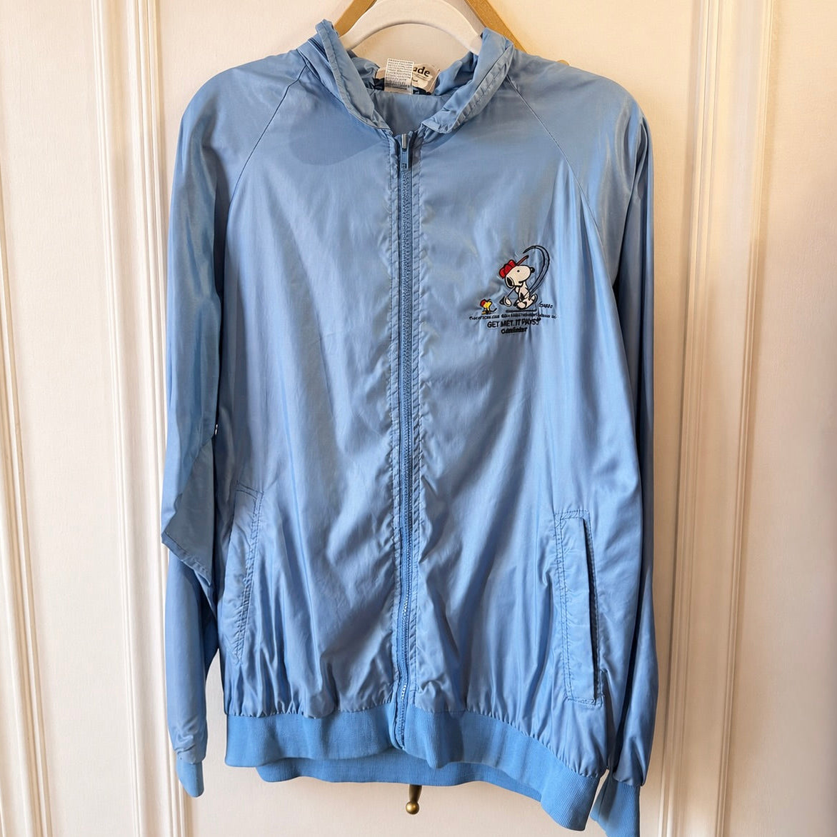 A light blue zip-up hooded jacket hanging on a wooden hanger against a cream-colored paneled door. The jacket features a small embroidered cartoon character on the left chest with text underneath.