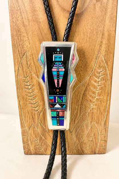 A geometric inlay bolo tie necklace with a black braided cord and a central rectangular pendant featuring colorful Southwestern-style geometric patterns on a black background, displayed against a carved wooden block.