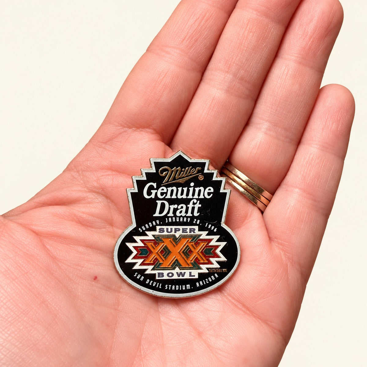 A close-up of a hand holding a Miller Genuine Draft Super Bowl LIX commemorative pin. The pin features a black and white design with orange and red accents, and is shown against a plain light beige background.