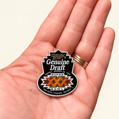 A close-up of a hand holding a Miller Genuine Draft Super Bowl LIX commemorative pin. The pin features a black and white design with orange and red accents, and is shown against a plain light beige background.