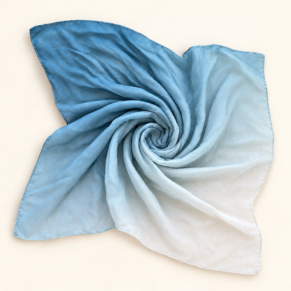 A square scarf with a soft ombré gradient transitioning from light blue to white, artistically arranged in a spiral on a warm off-white background.