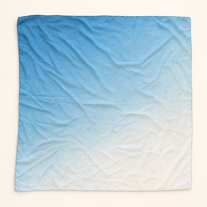 A square fabric scarf with a smooth blue-to-white gradient ombre design, laid flat on a light cream background.