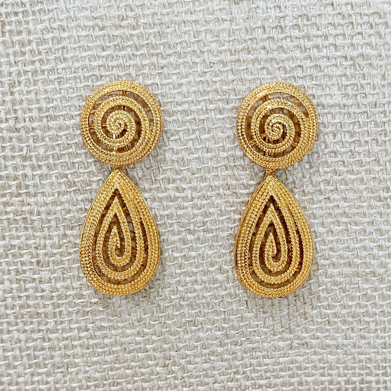 A pair of gold-toned drop earrings with spiral-shaped studs and teardrop-shaped dangling elements, displayed on a textured beige woven fabric background.