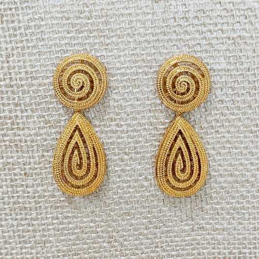 A pair of gold-toned drop earrings with spiral-shaped studs and teardrop-shaped dangling elements, displayed on a textured beige woven fabric background.