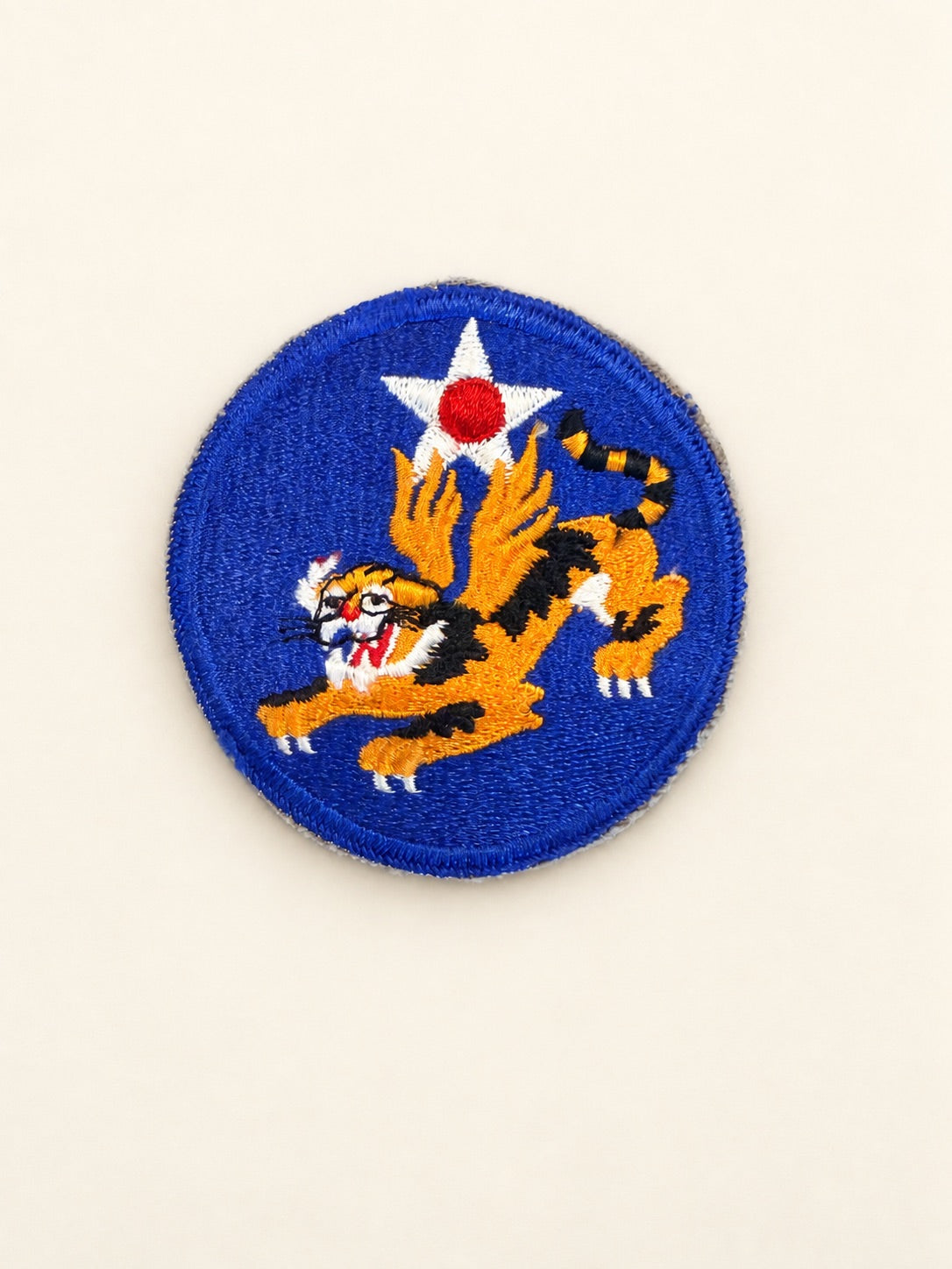 A circular embroidered patch featuring a blue background with a white star containing a red center, above a stylized orange and black tiger with white details, on a plain off-white background.