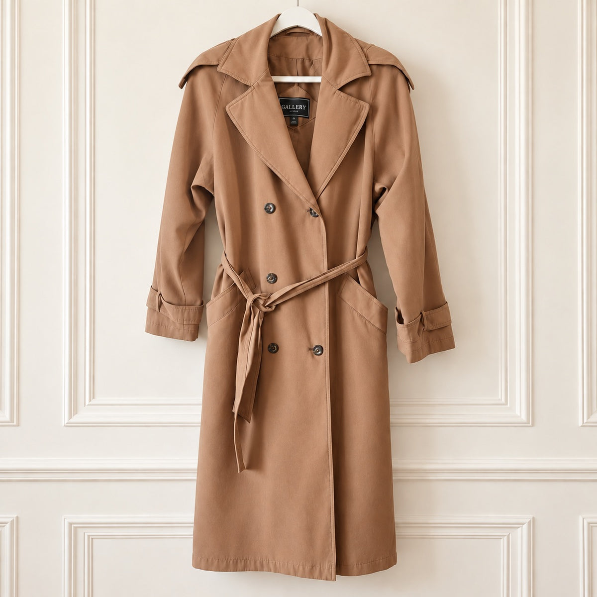 A classic double-breasted trench coat in camel brown, hanging on a white hanger against a white paneled wall. The coat features a matching waist tie, wide lapels, and button closures.