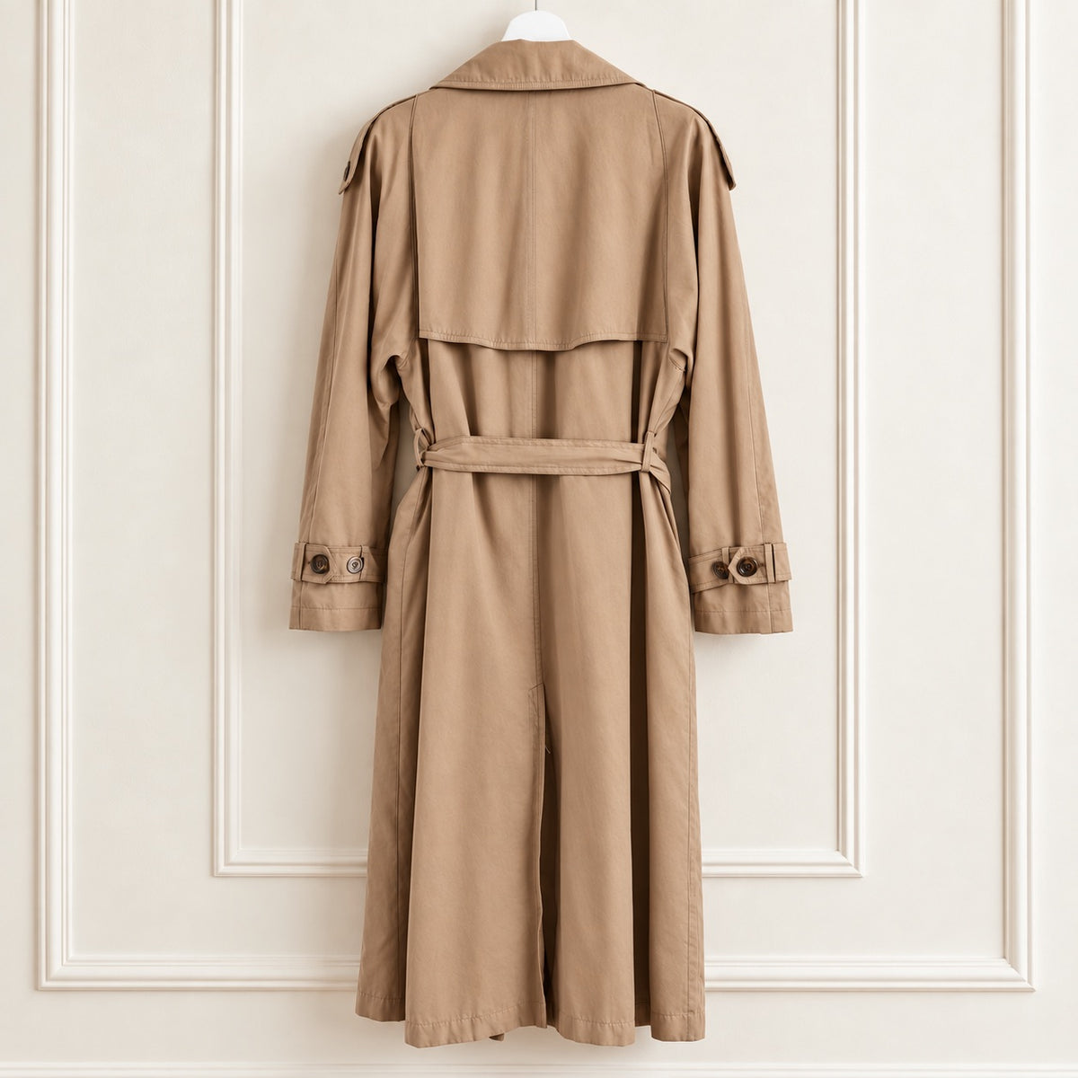 A classic trench coat in a warm camel brown color, hanging on a white hanger against a white paneled wall. The coat features a belted waist, long sleeves with buttoned cuffs, and shoulder epaulets.