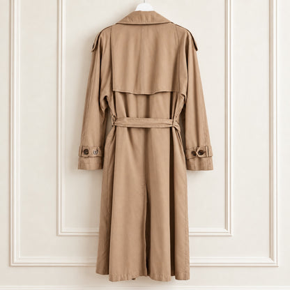 A classic trench coat in a warm camel brown color, hanging on a white hanger against a white paneled wall. The coat features a belted waist, long sleeves with buttoned cuffs, and shoulder epaulets.