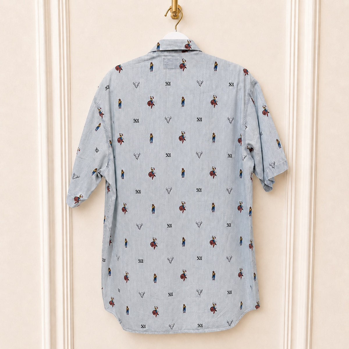 A light blue chambray short-sleeve button-up shirt with a repeating print of small cowboy figures and '501' text, hanging on a white hanger against a cream-colored paneled wall.