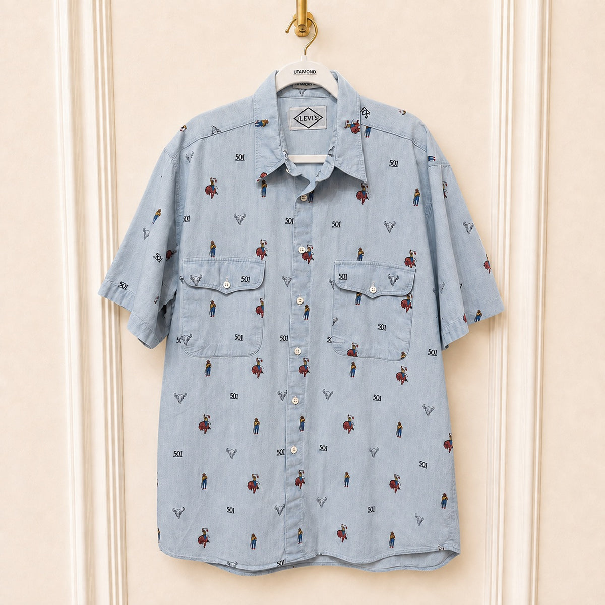 A light blue denim short-sleeve button-up shirt with an all-over print of small cartoon figures and the word '501' motifs, hanging on a white hanger against a cream-colored paneled wall.