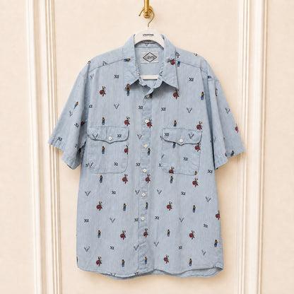 A light blue denim short-sleeve button-up shirt with an all-over print of small cartoon figures and the word '501' motifs, hanging on a white hanger against a cream-colored paneled wall.