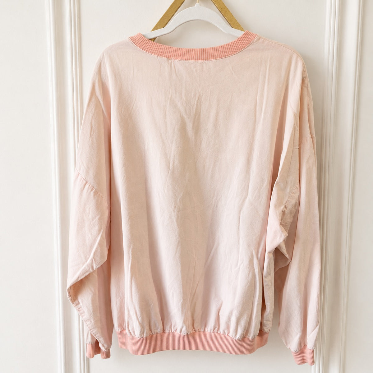 A light pink long-sleeve sweatshirt with ribbed crew neck, cuffs, and hem, hanging on a wooden hanger against a white paneled door.
