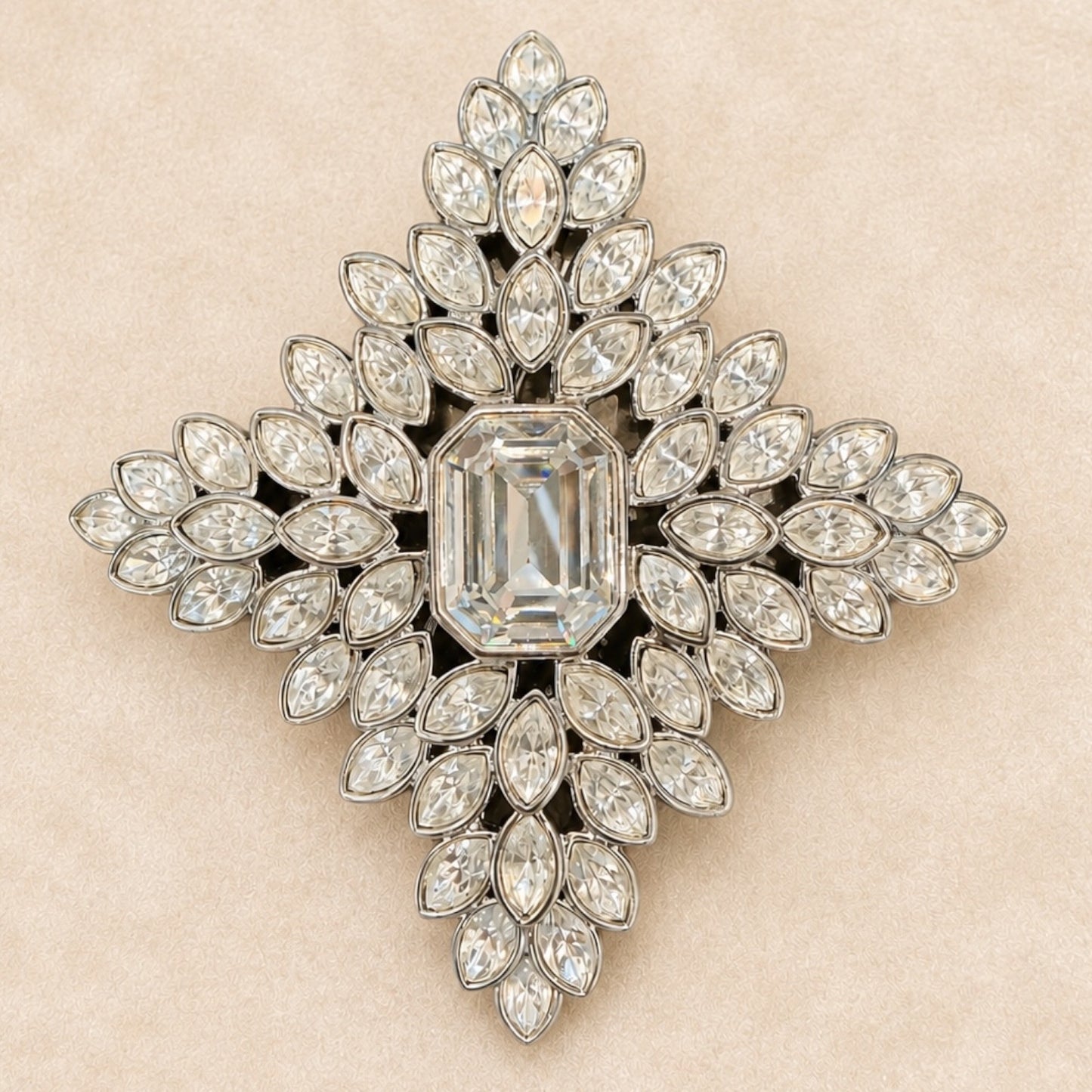 A vintage-style diamond brooch in a symmetrical cross or starburst shape, featuring a central emerald-cut diamond surrounded by multiple rows of marquise and round diamonds set in silver-toned metal, displayed on a textured beige background.