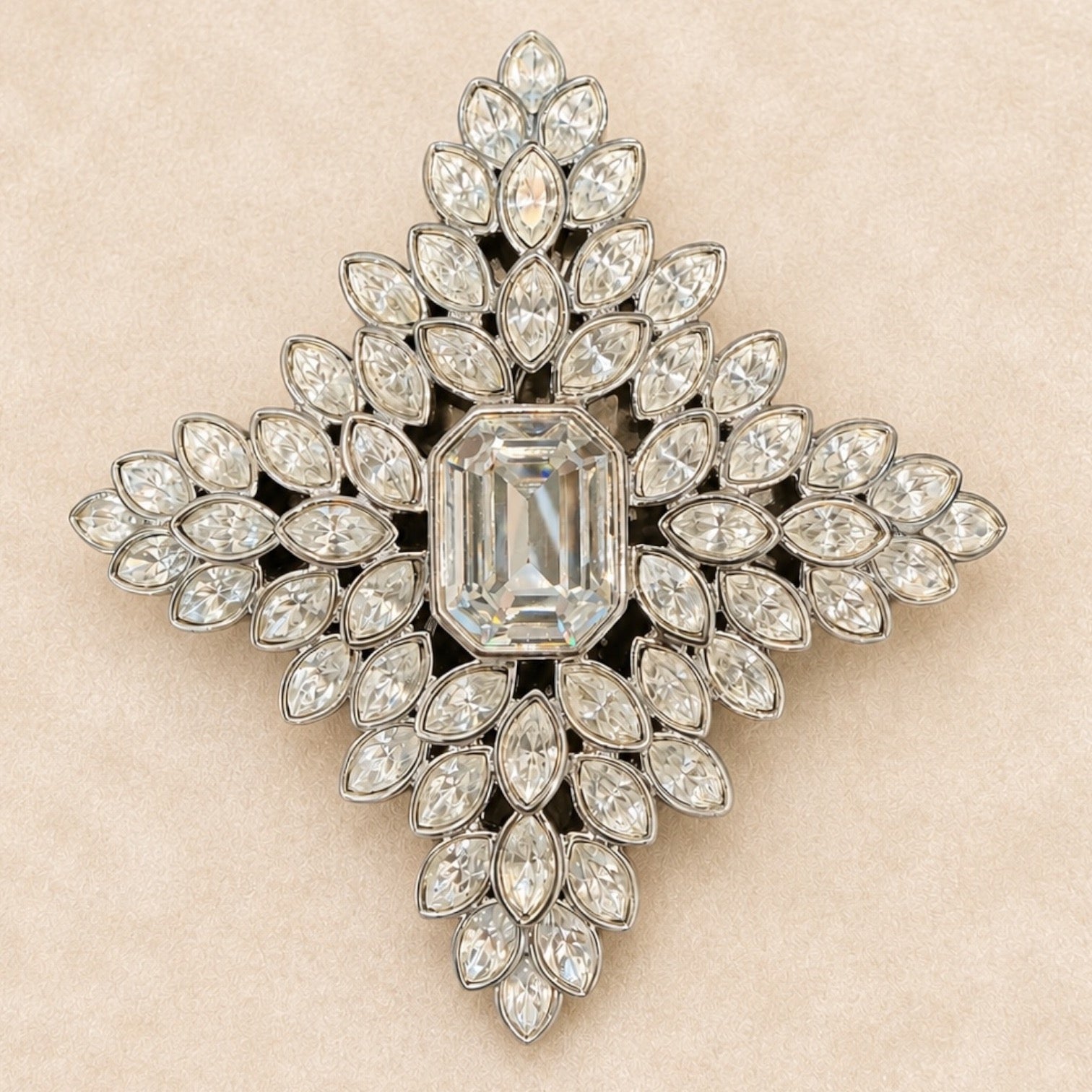 A vintage-style diamond brooch in a symmetrical cross or starburst shape, featuring a central emerald-cut diamond surrounded by multiple rows of marquise and round diamonds set in silver-toned metal, displayed on a textured beige background.