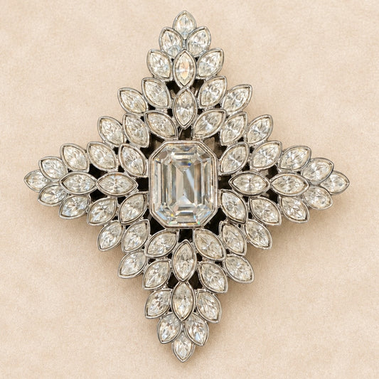 A vintage-style diamond brooch in a symmetrical cross or starburst shape, featuring a central emerald-cut diamond surrounded by multiple rows of marquise and round diamonds set in silver-toned metal, displayed on a textured beige background.