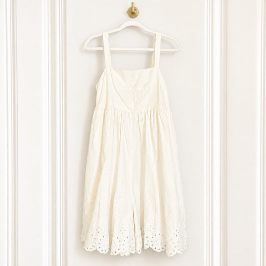 An off-white eyelet midi dress with thin shoulder straps hangs on a white plastic hanger from a gold hook against a white paneled door.