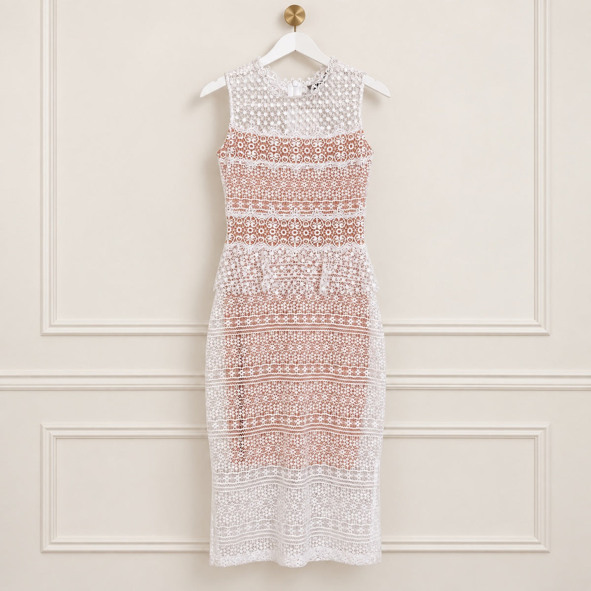 A sleeveless white crochet lace dress with a peach-colored patterned bodice, hanging on a white hanger against a cream-colored paneled wall.
