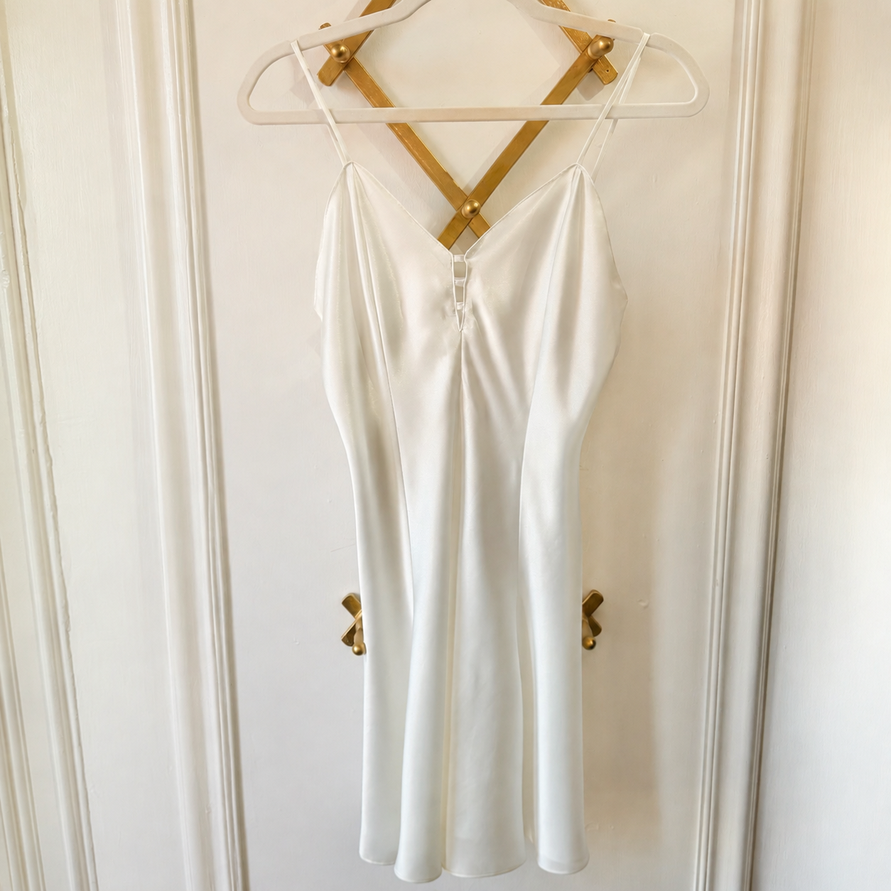 A white satin slip dress with thin spaghetti straps and a front zipper, hanging on a white hanger with gold-colored cross-back straps against a white paneled door.