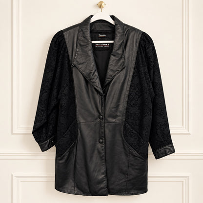 A black leather and jacquard jacket by Wilsons of London, hanging on a white paneled wall. The jacket features a smooth eather front panel, black patterned brocade sleeves, and a notched collar.