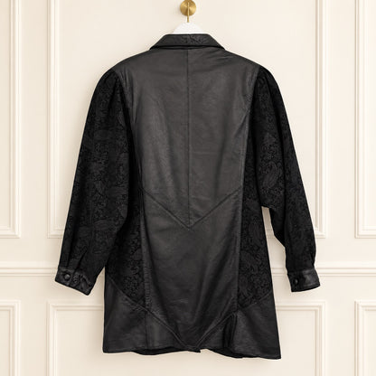 A black long-sleeve shirt jacket with textured jacquard patterned sleeves, hanging on a gold hanger against a white paneled wall.
