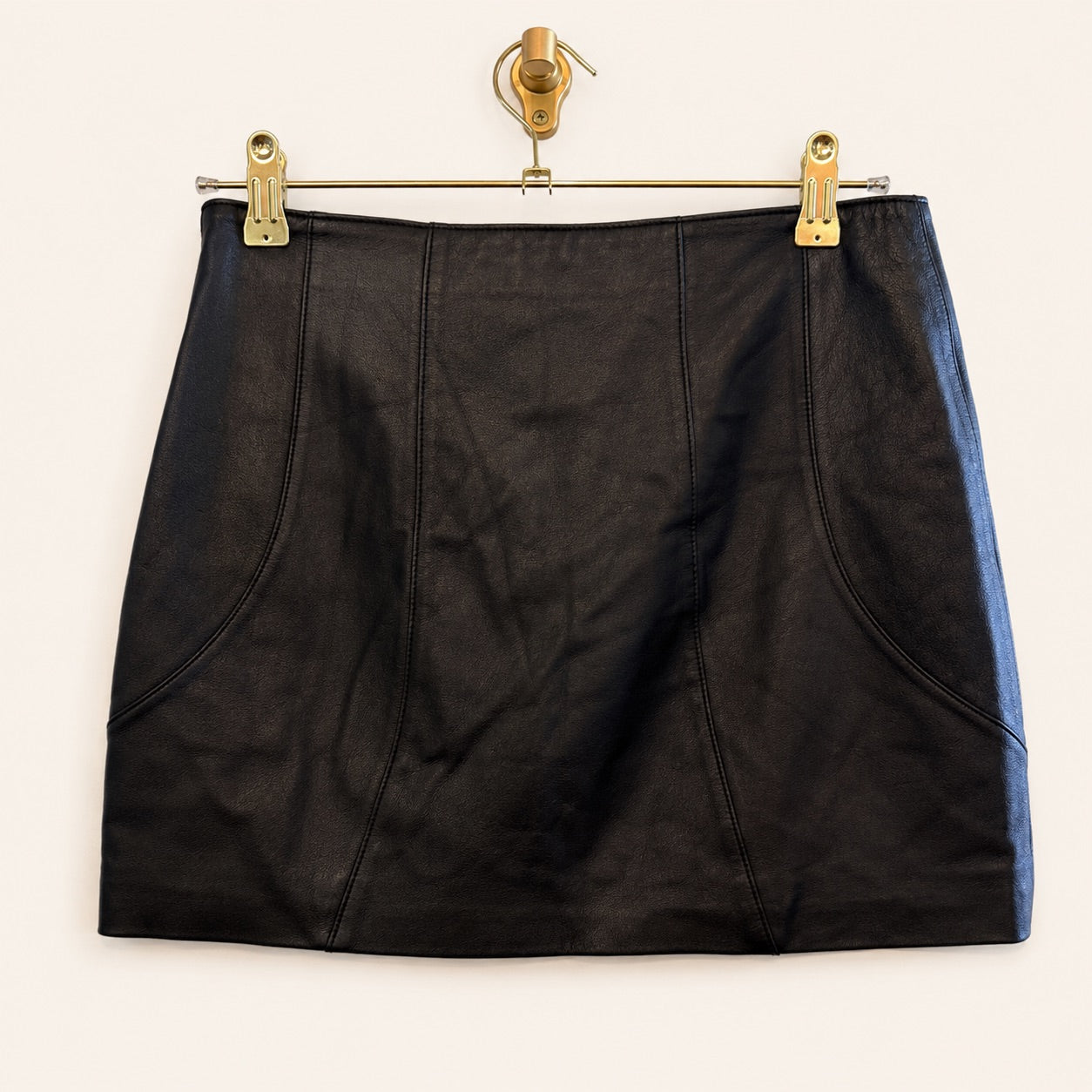 A black leather mini skirt, hanging from a gold-colored metal hanger with clips against a plain off-white background.