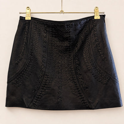 A black leather mini skirt with vertical perforated and braided cutout patterns, hanging from gold metal clips on a hanger against a plain off-white background.