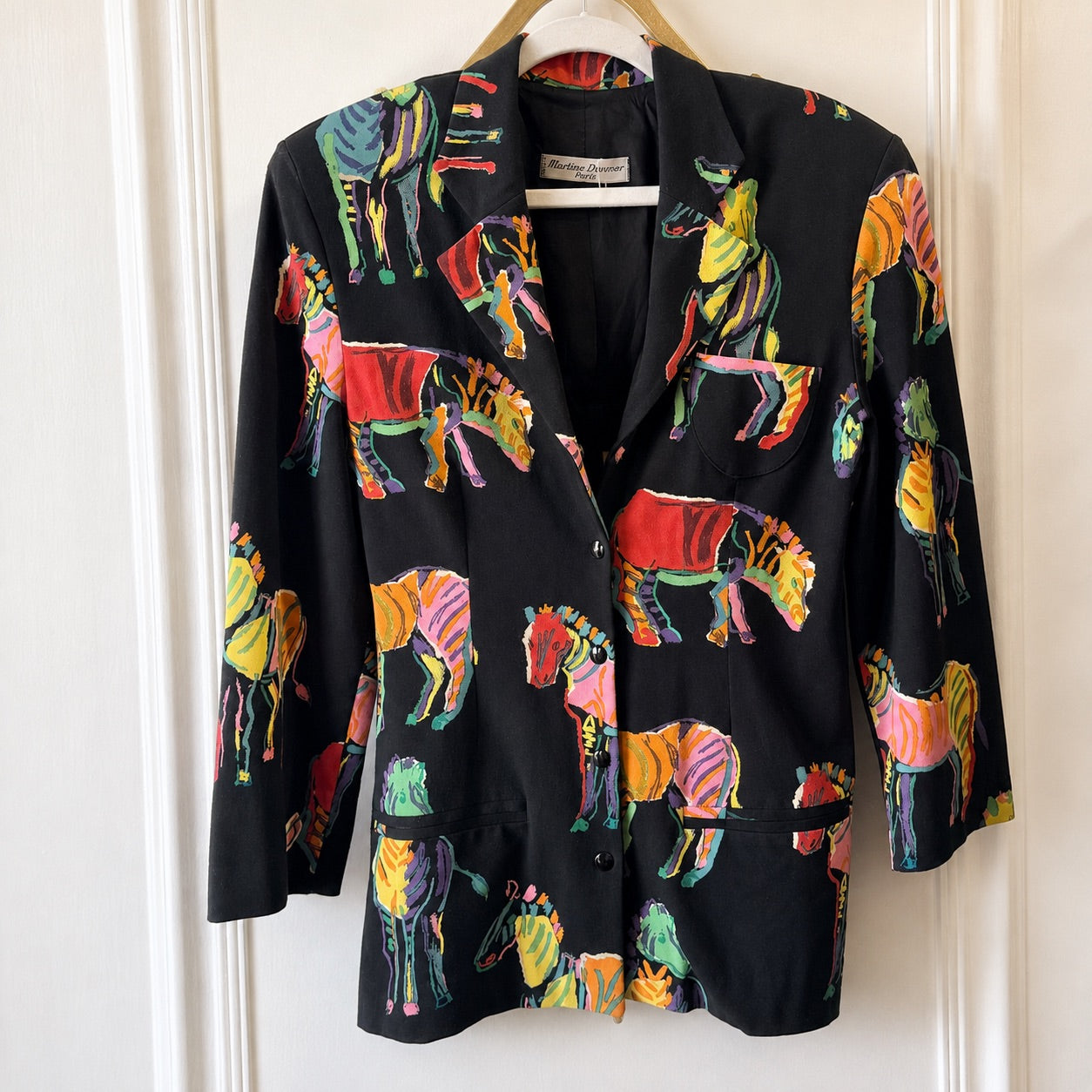 A black blazer jacket featuring a vibrant, multicolored zebra print, hanging on a wooden hanger against a white paneled door.