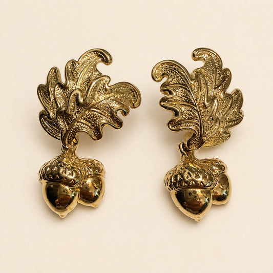 A pair of gold-toned drop earrings featuring textured oak leaves and acorns, displayed on a light beige background.
