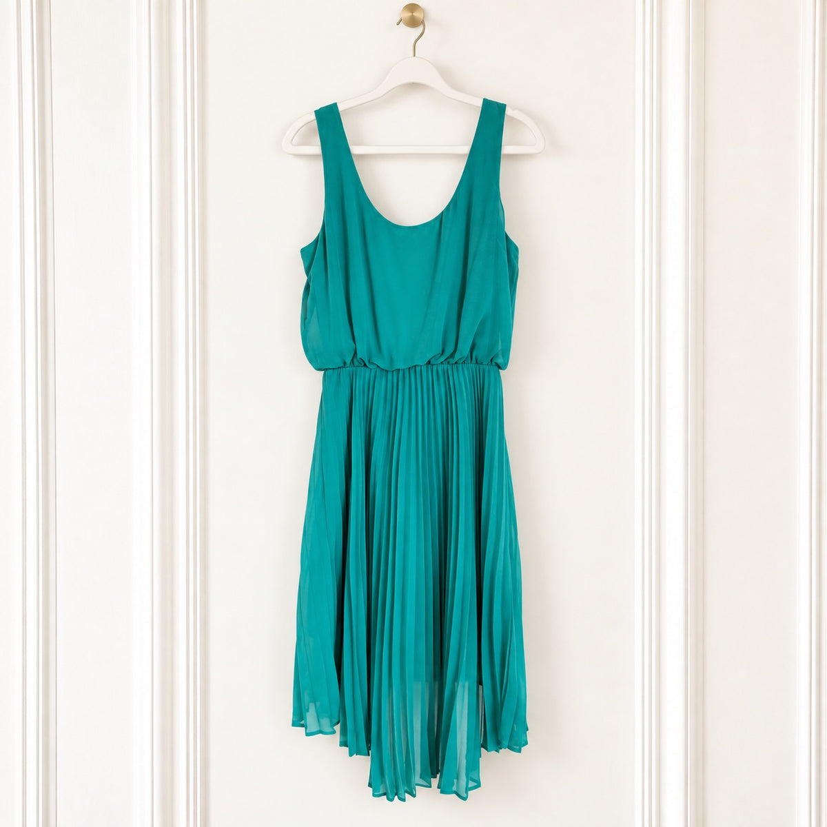 A sleeveless teal green dress with a scoop neckline and a pleated skirt, hanging on a white wooden hanger against a white paneled wall.