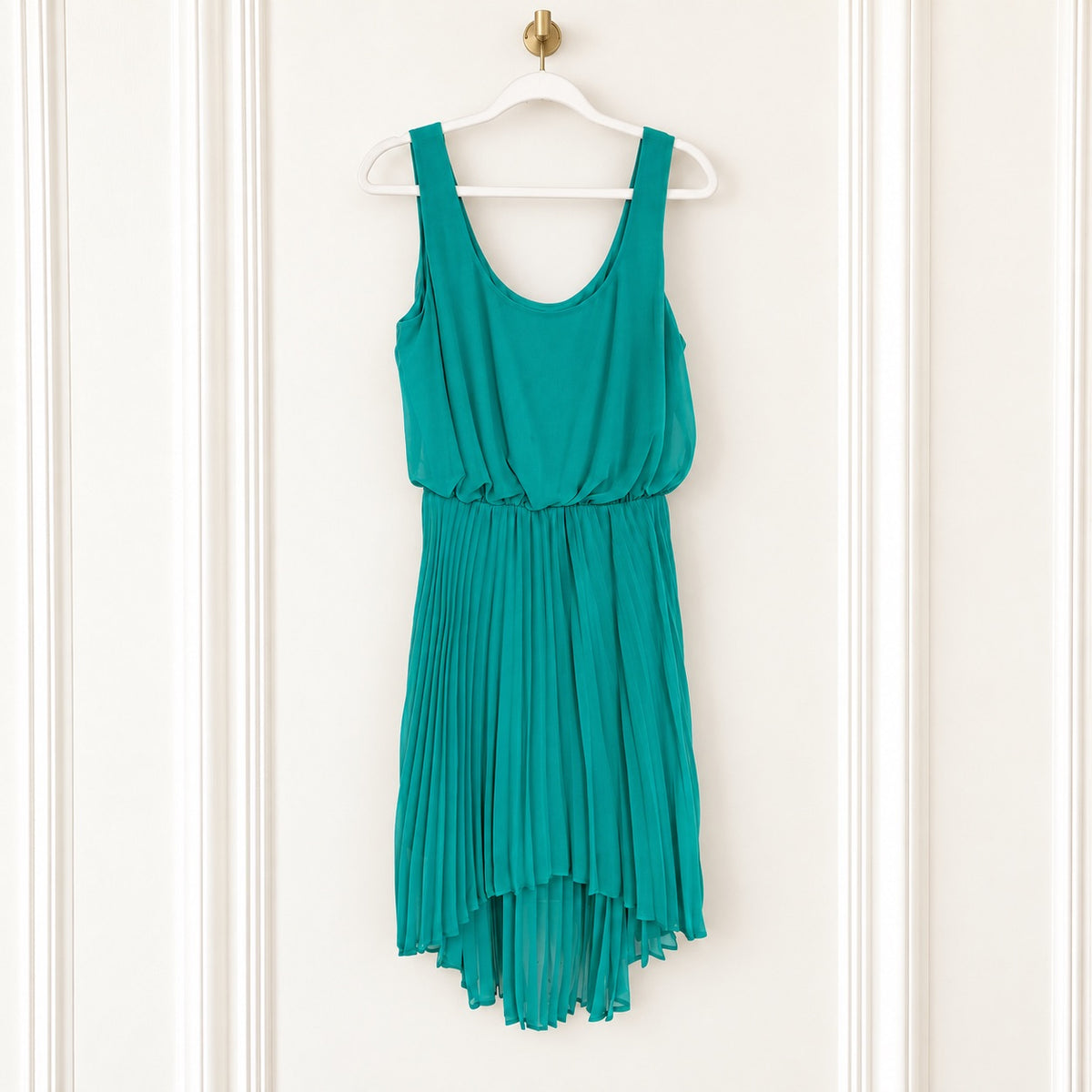 A teal green sleeveless dress with a pleated skirt, hanging on a white hanger against a white paneled door.