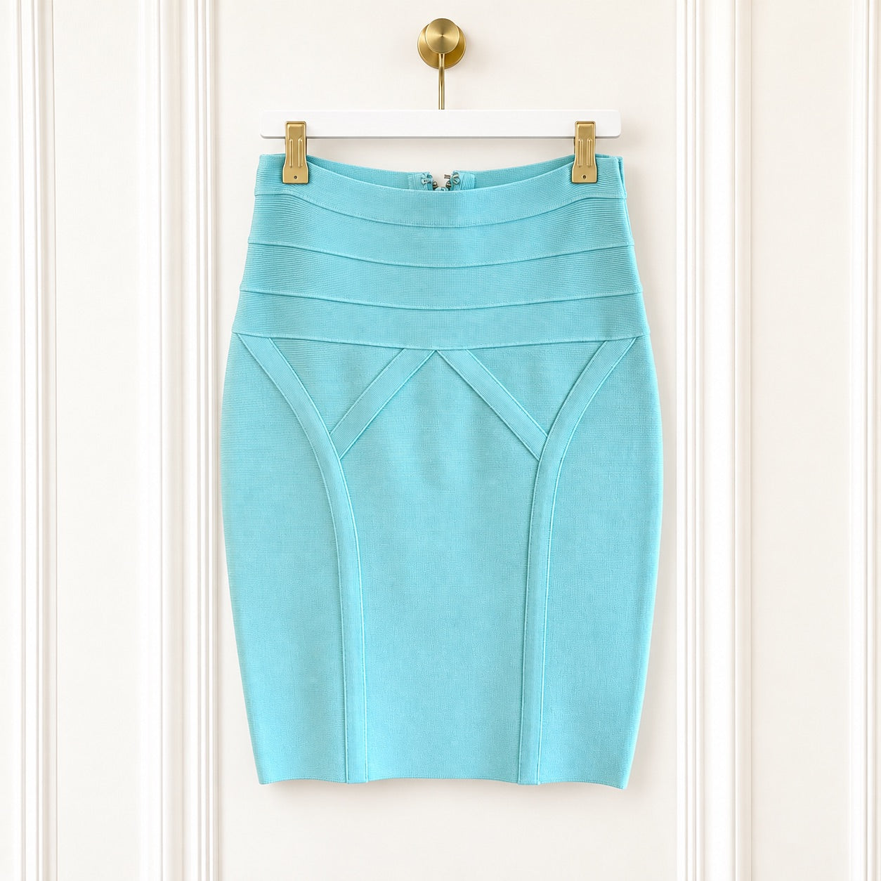 A light blue pencil skirt with structured paneling and diagonal seam details, hanging from gold clips on a white hanger against a white paneled door.