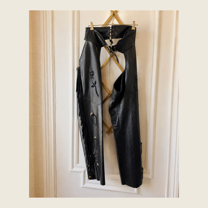 Black leather cropped pants with fringe, floral embroidery, and side grommets, hanging on a gold metal stand against a cream paneled wall.