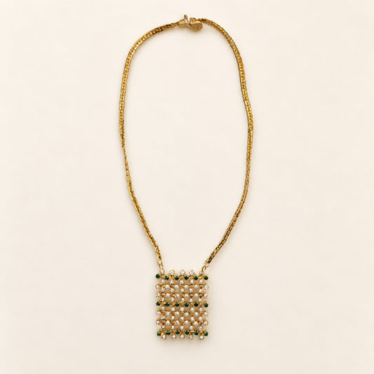 A gold chain necklace with a square pendant made of rows of small cream pearls and green gemstone accents, displayed on a light cream background.