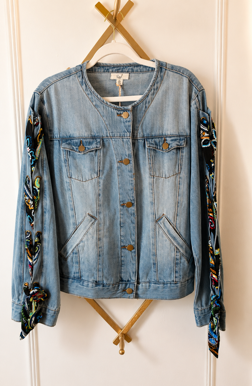 A light blue denim jacket with colorful patterned fabric draped over the sleeves, hanging on a wooden hanger against a white paneled wall.