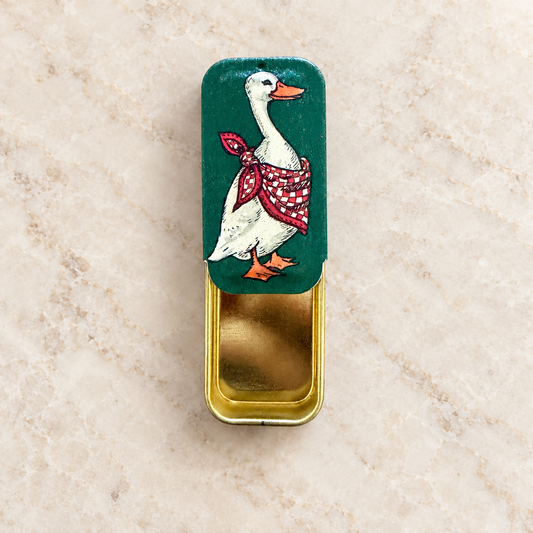 A small rectangular metal tin with a hinged lid, featuring a painted white goose wearing a red-and-white checkered scarf on a dark green lid. The tin is open to reveal a gold-toned interior, set on a light beige stone surface.