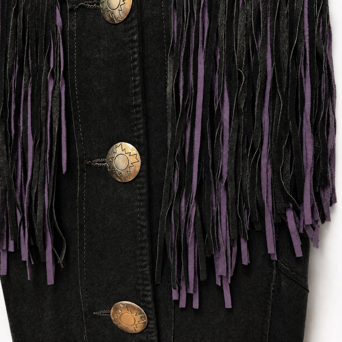 Close-up of a black denim jacket featuring long black and purple fringe trim and three bronze-toned decorative buttons.