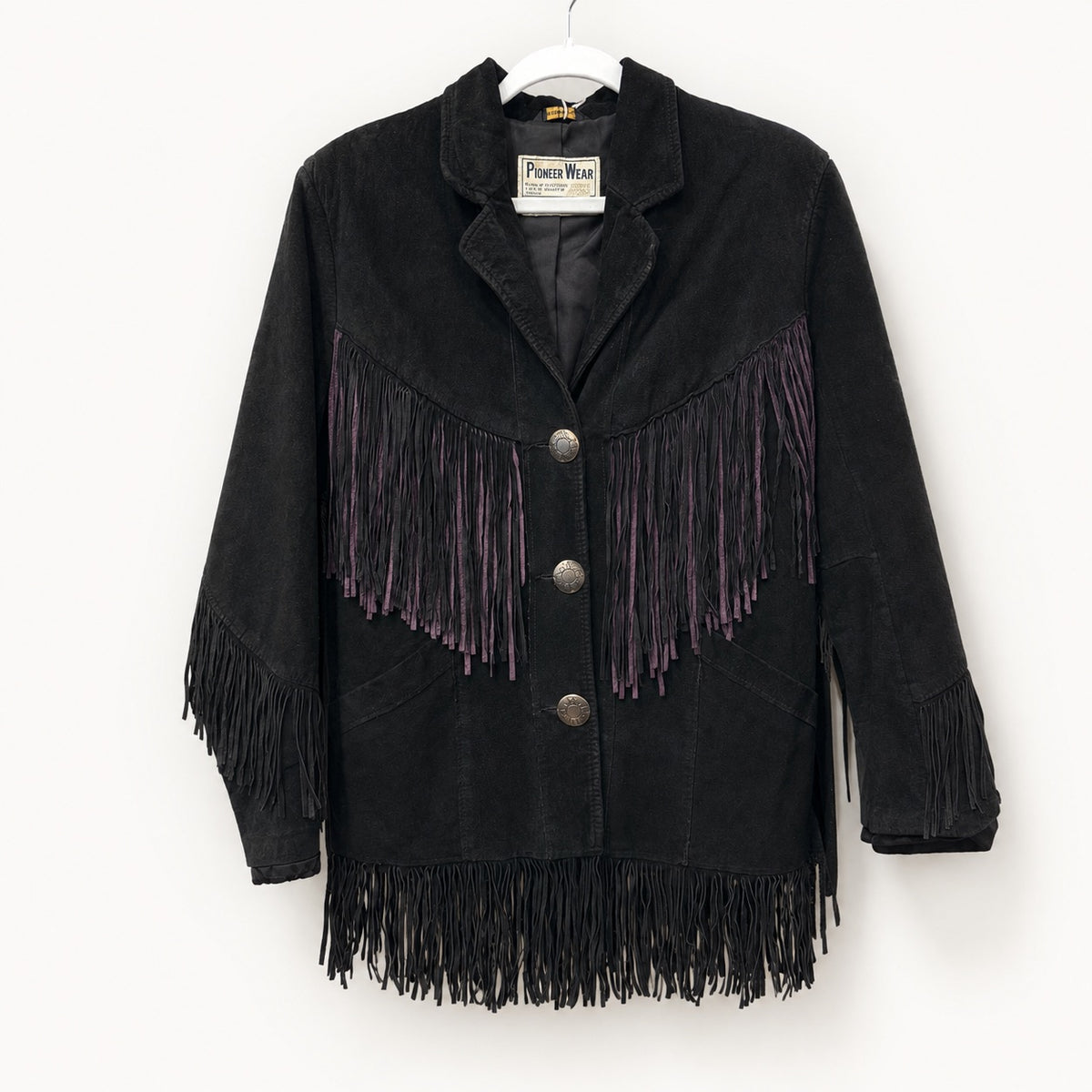 A black suede fringe jacket with purple fringe accents on the chest, hanging on a white hanger against a plain white wall. The brand label reads 'Pierce Wear'.