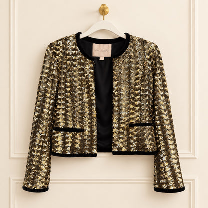 A cropped, long-sleeve sequined jacket in gold and black, hanging on a white hanger with a gold hook against a cream-colored paneled wall. The jacket features a black satin lining, black trim along the collar, pockets, and cuffs, and a visible 'marie h' brand label at the neckline.