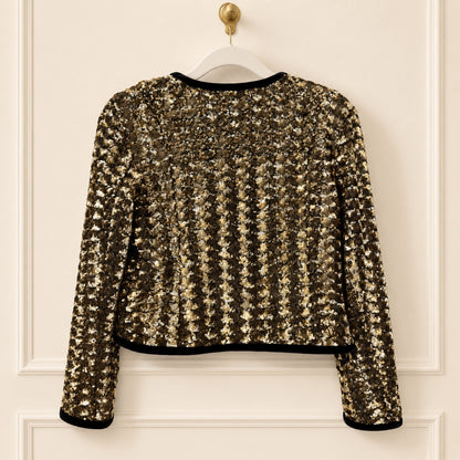 A cropped long-sleeve sequin jacket in a metallic gold and bronze shimmer, featuring black trim at the collar, hem, and cuffs, hanging on a white hanger against a cream-colored paneled door.