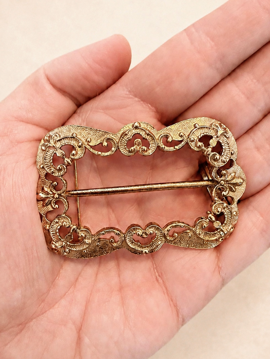 An ornate vintage-style gold-tone metal brooch with a decorative filigree border, held in the palm of a hand against a light beige background.