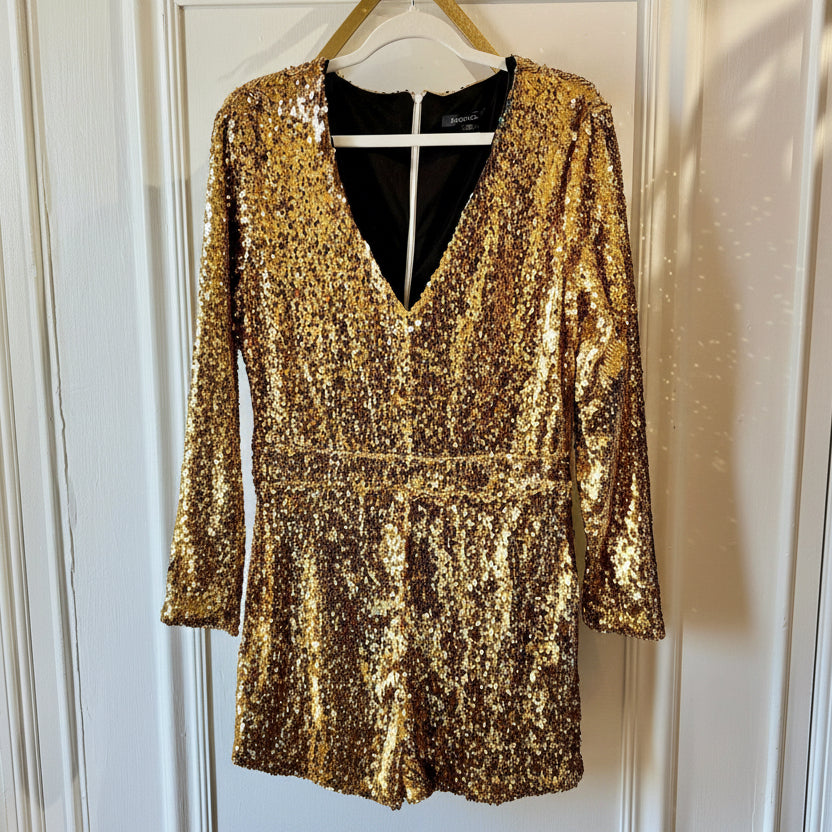 A gold sequined long-sleeve mini dress with a deep V-neckline, hanging on a wooden hanger against a white paneled door.