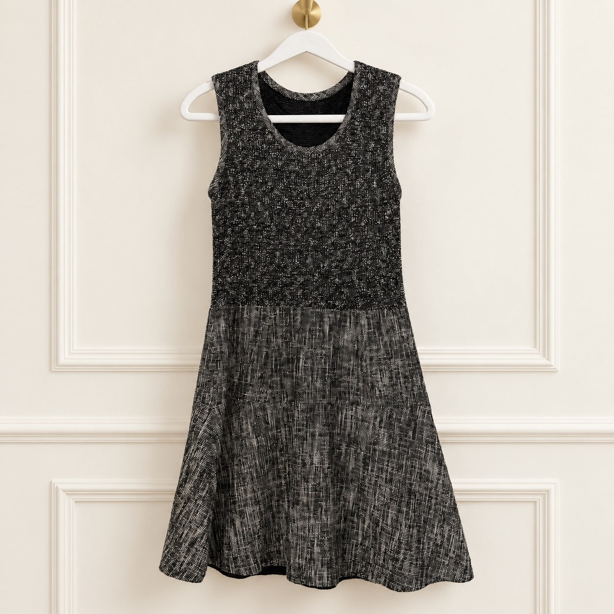 A sleeveless black and gray textured dress hanging on a white hanger against a cream-colored paneled wall.