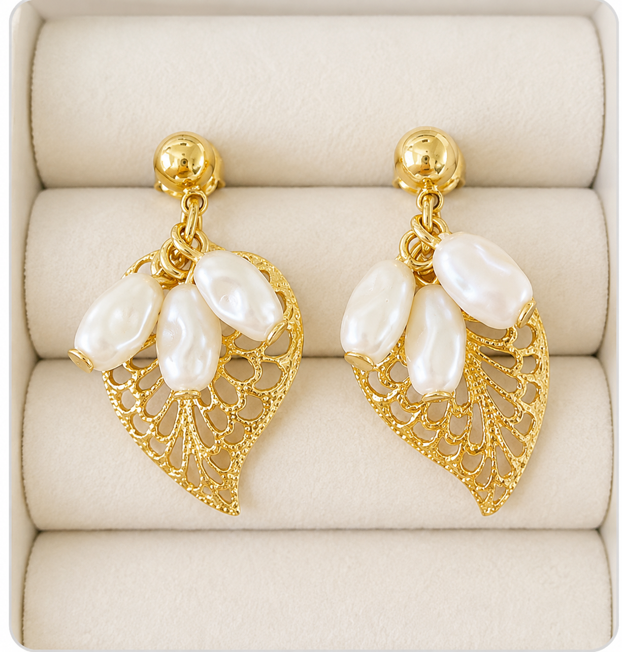 A pair of gold leaf drop earrings with three white baroque pearls, displayed on a beige jewelry box cushion.