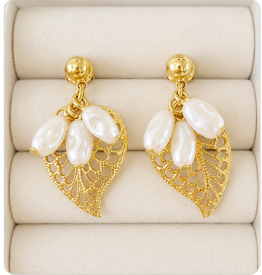 A pair of gold leaf drop earrings with three white baroque pearls, displayed on a beige jewelry box cushion.