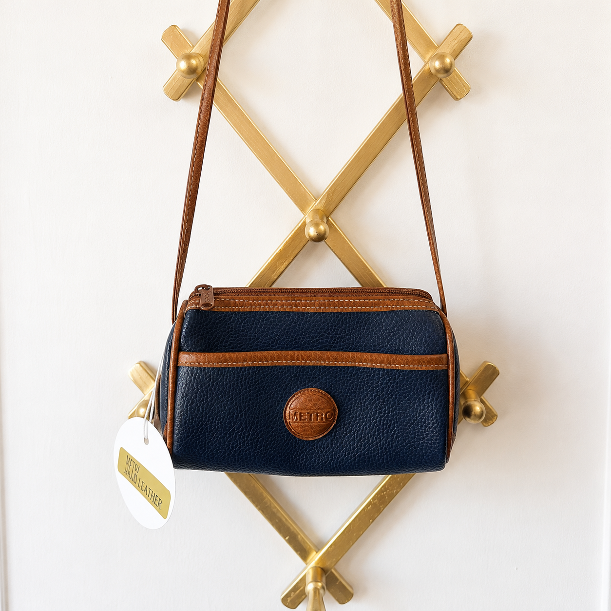 A navy blue pebbled leather crossbody bag with brown trim and a round brown logo patch, hanging from a gold geometric wall rack against a white wall.