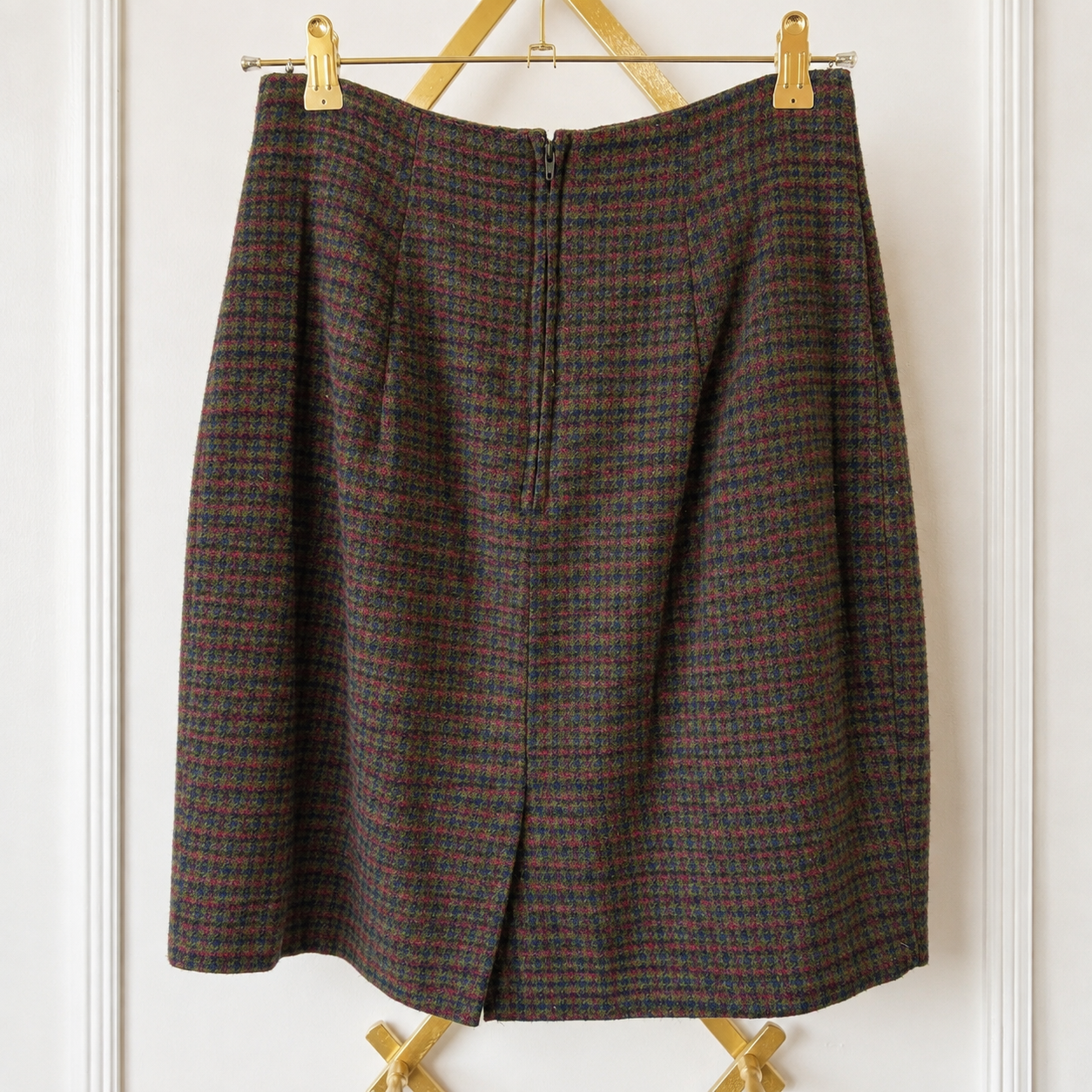 A vintage plaid skirt in brown, red, and black wool, hanging on a gold metal hanger against a white paneled wall.
