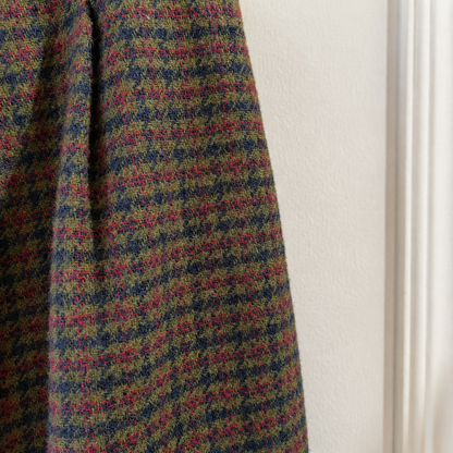 A close-up of a textured tweed fabric with a multicolor houndstooth pattern in olive green, navy blue, and magenta pink, hanging next to a white wall.