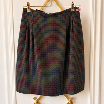 A pleated A-line skirt in a dark brown and red plaid tweed fabric, hanging from gold metal clips on a wooden hanger against a white paneled door.