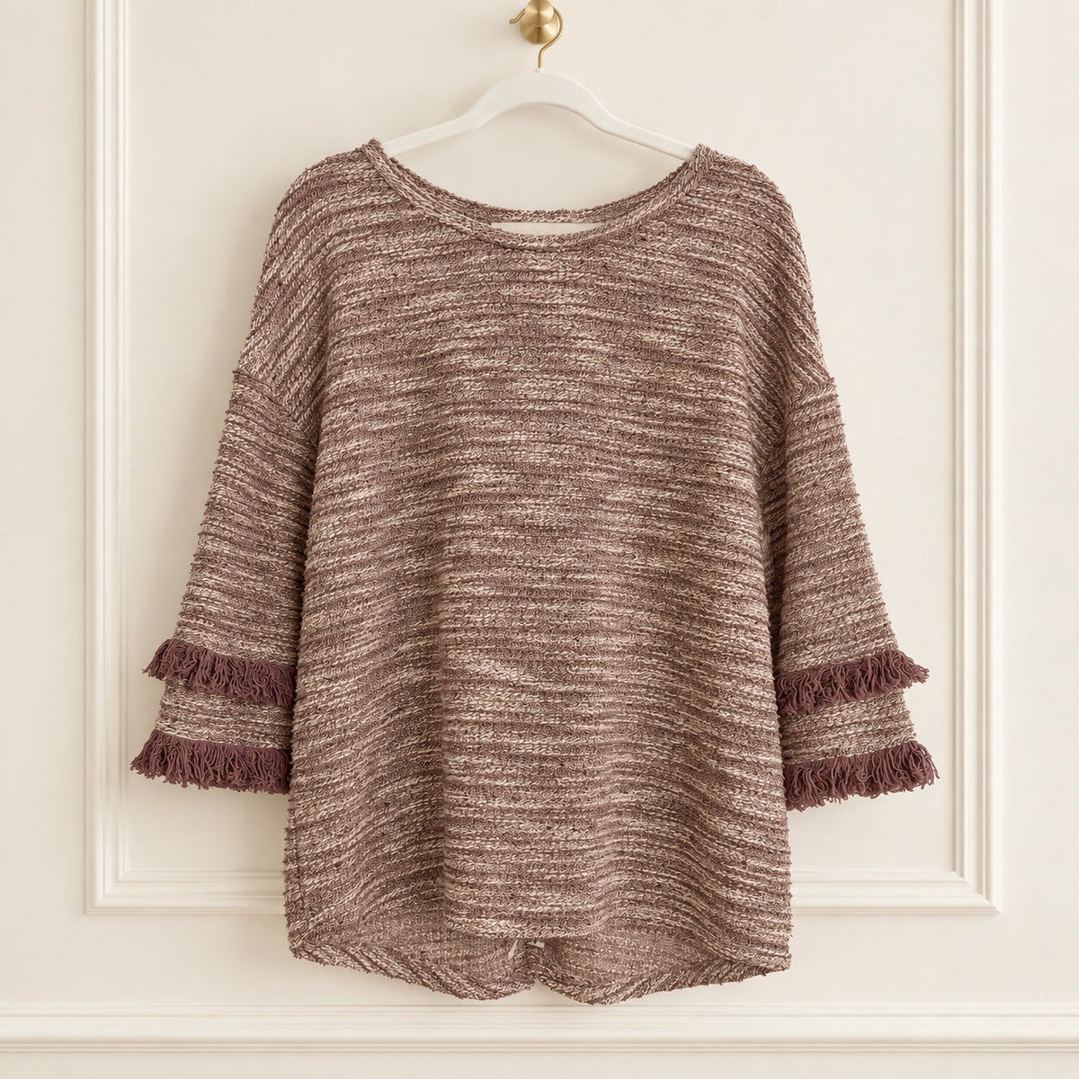 A brown and beige marled knit sweater with layered ruffle cuffs and fringe trim, hanging on a white hanger against an off-white paneled wall.